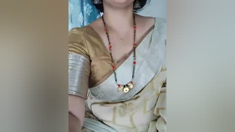Video of a South Asian woman wearing a golden sari with intricate patterns, adorned with red and black bead necklace and matching earrings, standing indoors against a blue floral curtain.