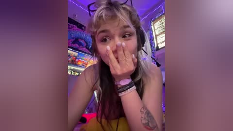 A young woman with long, dyed hair, wearing headphones, covers her face with her hand, in a brightly lit room with colorful decor.