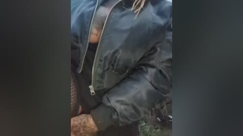 Media: Video of a person wearing a dark, shiny, waterproof jacket with a zippered front, standing outdoors in a forest. The jacket's texture is smooth and reflective, and the person's legs are partially visible, clad in dark, possibly camouflage-patterned pants.