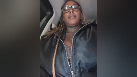 Media: A video of a middle-aged woman with braids and glasses, wearing a black leather jacket, seated in a car. The background is dark and blurry.