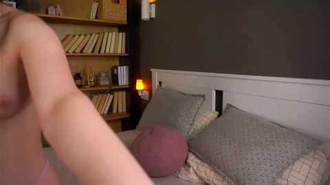 Video of a woman in a bed with gray pillows and beige blankets, partially naked with visible breasts. A beige shelf with books and a lamp is in the background.