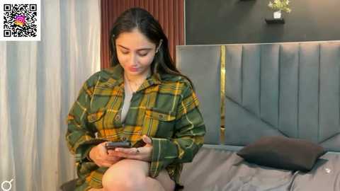 Video of a young woman with long brown hair, wearing a green plaid shirt, sitting on a gray bed, focused on her smartphone.