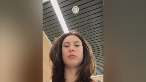 Video of a young woman with long, wavy brown hair, looking down, in a modern room with a gray ceiling and bright overhead lights.