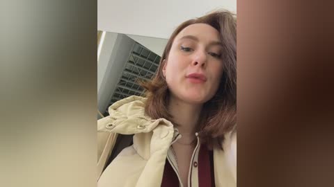 A video of a young woman with shoulder-length brown hair, wearing a beige jacket, blowing a kiss. The background features a blurred brick wall and a partially visible window.