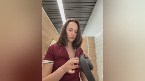 Video of a young woman with shoulder-length brown hair in a maroon shirt, adjusting a black microphone in a modern, minimalist restroom with light wood and white tiles.