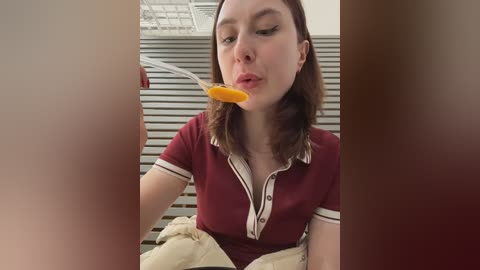 Video of a young Caucasian woman with shoulder-length brown hair, wearing a burgundy polo shirt with white trim, eating a spoonful of orange yogurt. Background includes a white shelving unit and beige fabric.