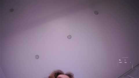 Video of a dimly lit room with a textured, pale purple ceiling featuring three circular light fixtures. A person's face is partially visible at the bottom, their hair disheveled.