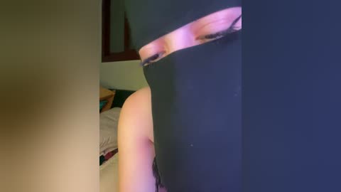 Media: A video of a person wearing a black mask, with a partial view of their face and a blurry bedroom background, featuring a bed and wall.