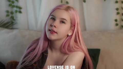 Video of a young, fair-skinned woman with long, straight pink hair, wearing a black top, sitting on a beige couch. She has a neutral expression. Background includes green plants and white curtains. Text at the bottom reads \"LOVENSE IS ON.\