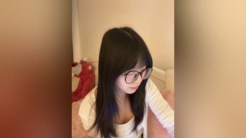 Video of an Asian woman with long black hair, wearing glasses and a white top, sitting on a pink bed with a stuffed animal.