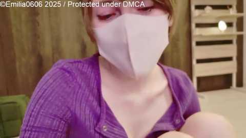 Video of a young woman with fair skin, short brown hair, and wearing a purple knit cardigan and a white surgical mask, holding a baby. Background features wooden walls and a white crib.