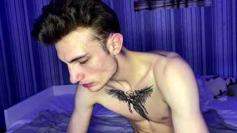 Video of a shirtless young man with fair skin, dark hair, and a prominent black angel wings tattoo on his chest. He's indoors, kneeling on a bed with white sheets, against a blue striped wall.