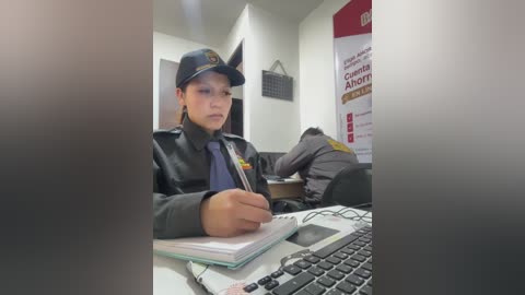 A video of a woman in a police uniform with a black cap, writing in a notebook at a desk. Background shows another officer and a poster.