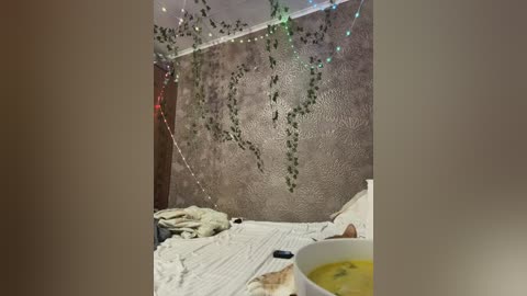 A video of a cozy bedroom with a beige textured wall adorned with green string lights and hanging vines. The bed, with a white sheet and pillow, is disheveled, and a bowl of yellow soup sits on a bedside table.