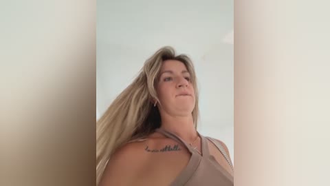 Video of a blonde woman with long hair, wearing a beige top, looking upward. A tattoo reads \"Be still\" on her right shoulder. Background is a plain, light-colored wall.