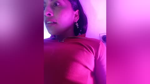 Video of a light-skinned woman with shoulder-length dark hair, wearing a pink top and dangling earrings, taken indoors with pink lighting, showing her face partially obscured by hands.