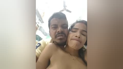 A video captures a South Asian couple in an intimate moment, with the man behind the woman, both topless. The background features a ceiling fan and a blurred kitchen setting.