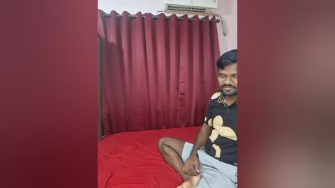Video of a South Asian man with a beard, wearing a black t-shirt with yellow patterns and grey shorts, sitting on a red bed in a dimly lit room with red curtains and an air conditioning unit.