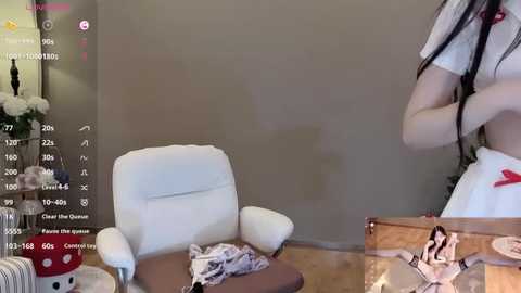 Video of a young woman with long black hair in a white nurse outfit, standing in a cozy, minimalist room with a white chair, wooden floor, and floral decorations.