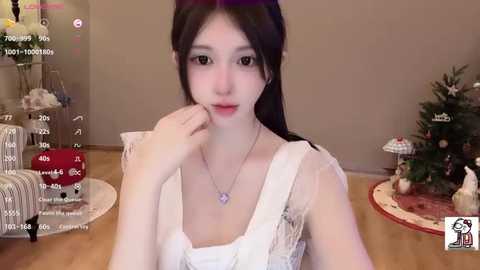 Media: Video of a young East Asian woman with pale skin, long black hair, and brown eyes, wearing a white sheer dress with a heart pendant necklace. Background features a Christmas tree, wrapped presents, and a festive table setting.