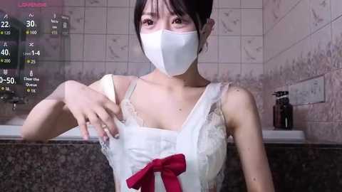 Video of an Asian woman with short dark hair, wearing a white lace dress, white mask, and red bow. Background shows a tiled bathroom with a soap dispenser.