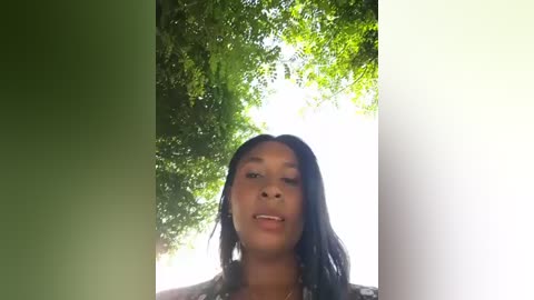 Video of a young Black woman with long, dark braided hair, wearing a patterned top, smiling against a bright, sunlit background with lush green trees.