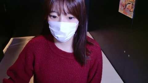 Video of an Asian woman with straight, shoulder-length brown hair, wearing a white surgical mask and a red knit sweater, standing indoors against a dark background with a colorful poster on the wall.