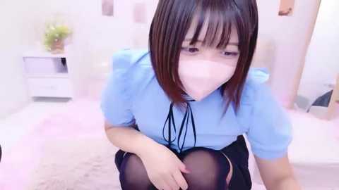 Video of an Asian woman with straight black hair, wearing a light blue blouse and black skirt, sitting on a bed with a pink blanket, wearing a face mask.