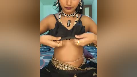 Video of a woman lifting a black crop top to reveal her medium-sized breasts. She wears a gold chain, hoop earrings, and a black sari with intricate gold embroidery. The background shows a bed with patterned sheets.