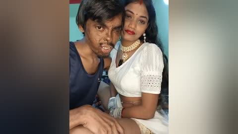 Video of an Indian couple in traditional attire: the man wears a blue shirt, and the woman, a white saree with gold jewelry. They sit closely, smiling. Background is blurred.