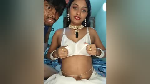 Video of a South Asian woman with medium skin tone, black hair, wearing a white bra and gold necklace, standing in a bedroom with a man behind her.