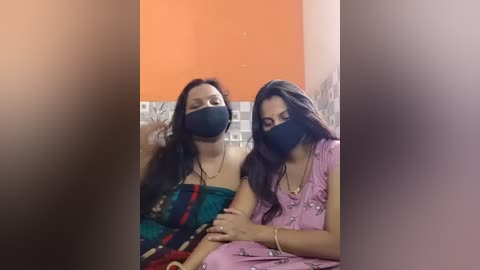 Video of two women with long black hair, wearing black masks and colorful saris, seated indoors against an orange wall with tiled background, one with a gold chain.