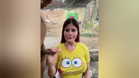 Video of a young woman with long red hair, wearing a yellow SpongeBob shirt, sitting on a bench outdoors. A man's erect penis is visible in the foreground.