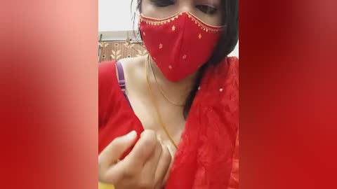 Video of a woman in a red sari and matching red face mask, adjusting her necklace. Background shows patterned fabric.