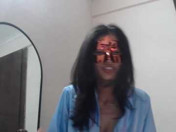 Video of a woman with long black hair, wearing reflective sunglasses and a blue shirt, standing in a room with white walls and a large mirror.