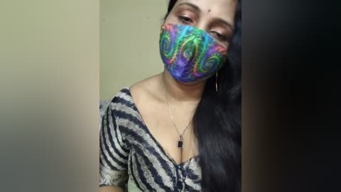 Video of a woman with medium skin tone and long black hair, wearing a face mask with vibrant, psychedelic spiral patterns, a black and white striped top, and a necklace.