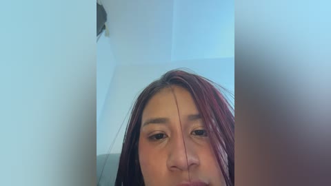 A close-up video of a young woman with medium brown skin and long, wet red hair, looking directly at the camera. She has a slight frown, and her eyes are partially obscured by her hair. The background is blurred, showing a white wall.
