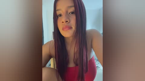 Media: A video of a young Black woman with long, straight, reddish-brown hair and medium skin tone, wearing a red strapless top, making a pouty face. The background is blurred, showing a white wall and a bed.