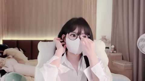 A video of an East Asian woman with long black hair and glasses wearing a white face mask, adjusting it on a bed with beige sheets, beige curtains, and a nightstand with a fan.