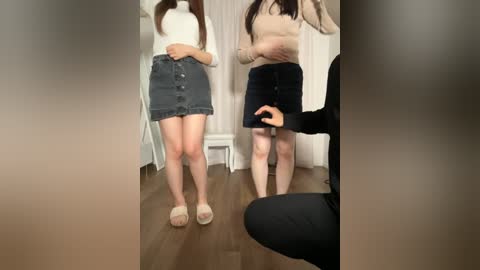 Media: Video of two women in casual attire; one in a beige sweater and dark denim skirt, the other in a black sweater and skirt, standing in a room with wooden floor and white curtains.