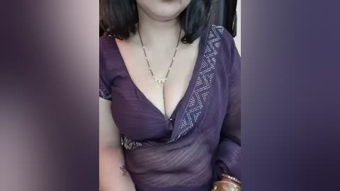 Video of a fair-skinned woman with shoulder-length black hair, wearing a deep purple, sheer blouse with white geometric patterns, revealing ample cleavage. She accessorizes with a gold necklace and a brown bracelet. Background is blurred, indoor setting.