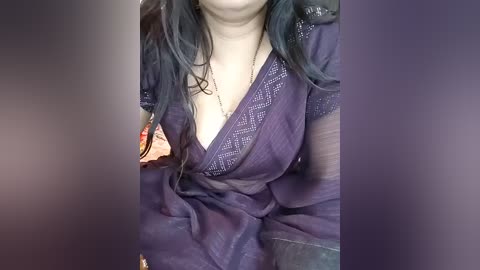 Video of a woman with long, wavy, black hair, wearing a deep purple, sheer dress with lace details, revealing cleavage, against a blurred background.