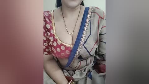 Video of a woman in a red and beige sari with intricate gold patterns, blue shoulder strap, and gold necklace, viewed from the shoulders down.