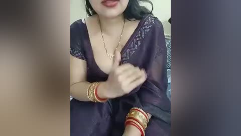 Video of a South Asian woman with fair skin, wearing a deep purple sari with intricate gold embroidery, gold bangles, and a gold necklace. She is seated indoors with a blurred background.