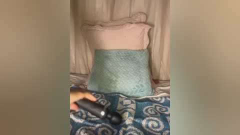 Video of a beige curtain backdrop with a blue and white patterned blanket, a teal pillow, and a person holding a black gaming controller.