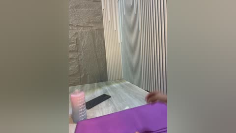 Video of a small, modern bathroom with a light grey brick wall, vertical white radiator, and a purple yoga mat on a light wooden floor. A person's hand is on the mat, and a pink bottle and black remote are nearby.