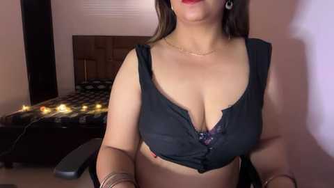 Video of a curvy woman with fair skin, dark hair, red lipstick, wearing a black lace bra and black pants, standing in a dimly lit bedroom with a checkered bed, string lights, and a wooden headboard.