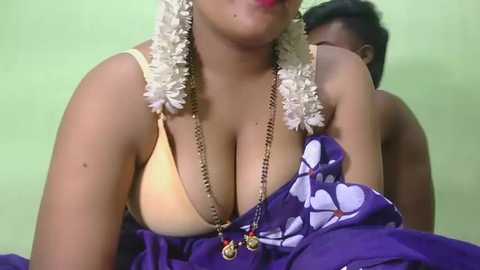 Video of a topless South Asian woman with medium skin tone, wearing a white garland, yellow bra, and purple saree with white floral print. Background is plain green wall.