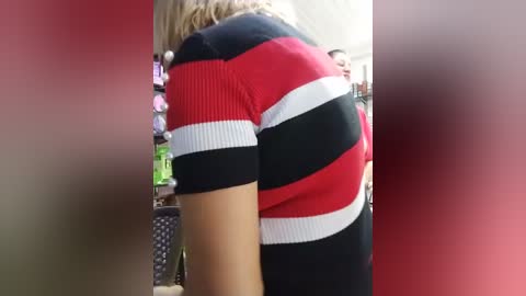 Video of a woman in a red and black striped top, her back turned, in a blurry background with a red wall and a green object.