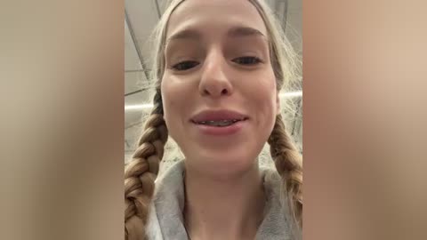 Video of a young Caucasian woman with light skin, blonde braided hair, and a slight smile, wearing a light gray shirt. Background shows an indoor setting with ceiling tiles.